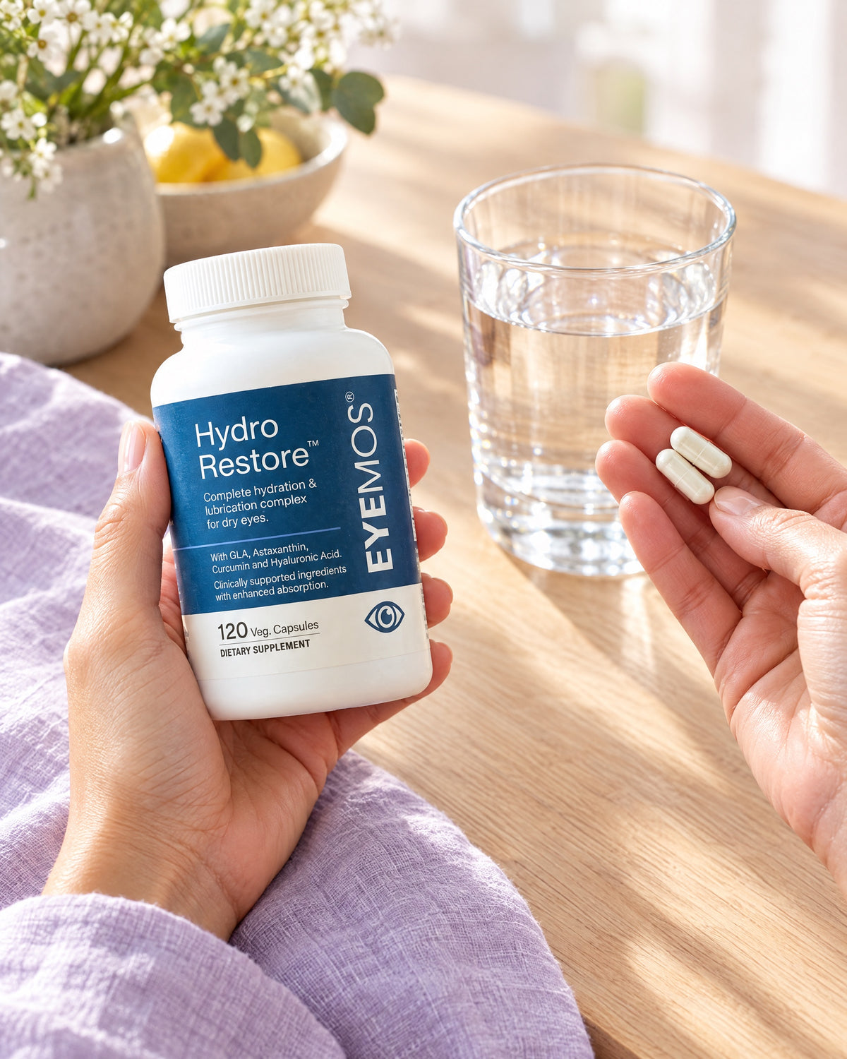 Hands holding a Hydro Restore bottle and two capsules beside a glass of water at a kitchen table